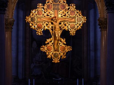 The image shows a large, ornate golden crucifix hanging from the ceiling inside a church, viewed through an archway with marble columns and an altar in the foreground.