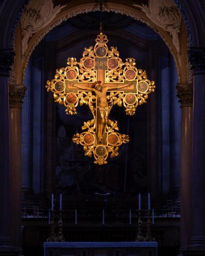 The image shows a large, ornate golden crucifix hanging from the ceiling inside a church, viewed through an archway with marble columns and an altar in the foreground.