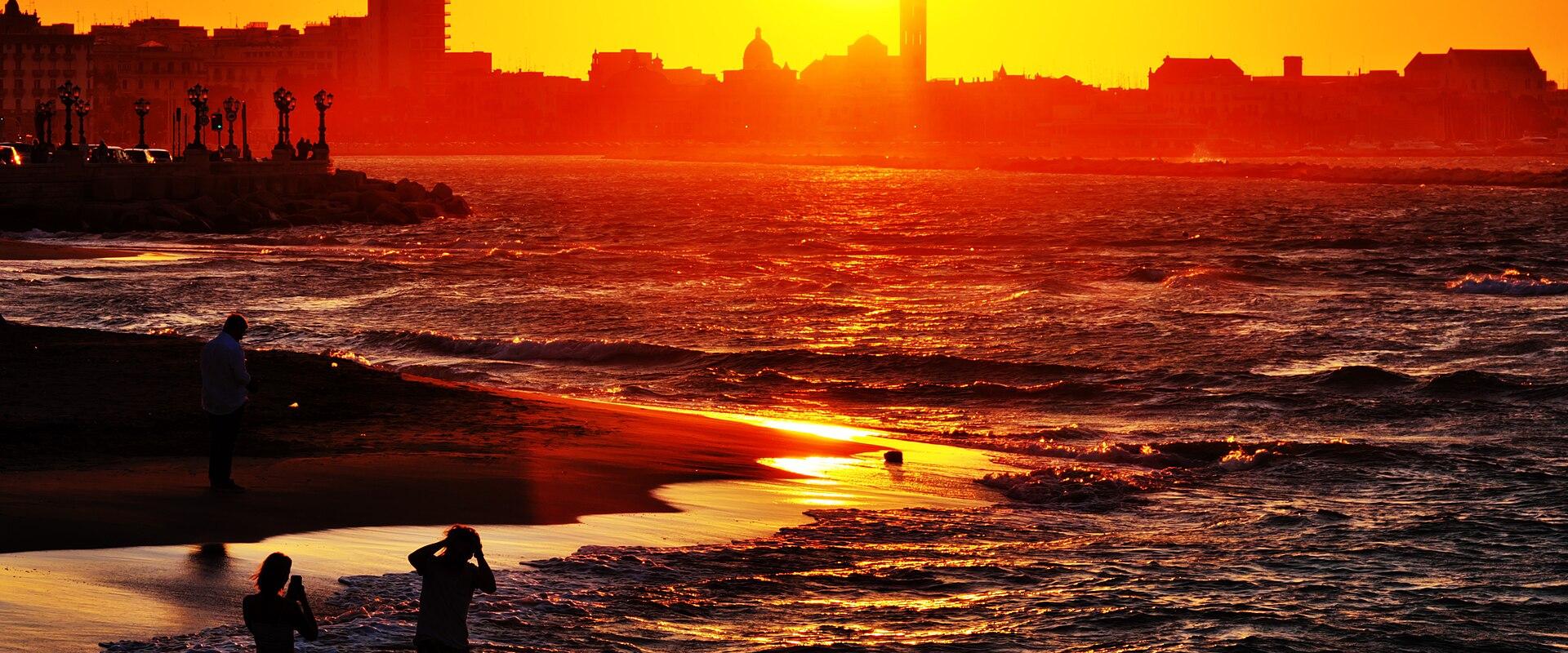 A vibrant sunset over a coastal city skyline with silhouetted figures on the beach in the foreground; a prominent church tower and domed buildings are visible along the waterfront.