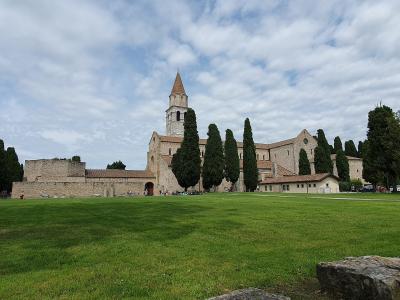 A wide-angle outdoor shot of a large, ancient stone basilica with a prominent bell tower and conical roof, set against a cloudy sky, with a manicured green lawn in the foreground and tall cypress trees lining the building; no visible signage or te