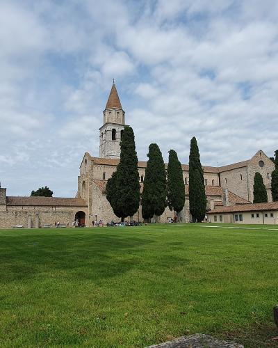 A wide-angle outdoor shot of a large, ancient stone basilica with a prominent bell tower and conical roof, set against a cloudy sky, with a manicured green lawn in the foreground and tall cypress trees lining the building; no visible signage or te