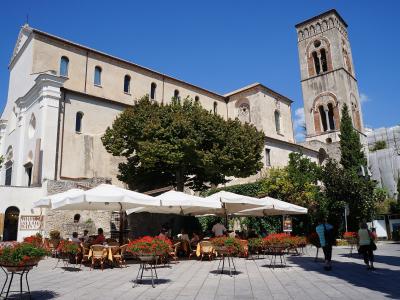 The image shows a large, historic stone cathedral with a prominent bell tower, situated behind an outdoor cafe with white umbrellas and potted red flowers under a clear blue sky.