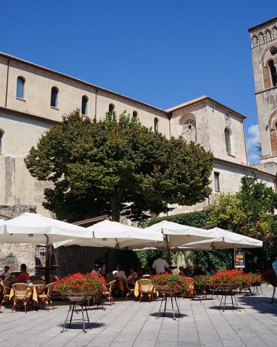 The image shows a large, historic stone cathedral with a prominent bell tower, situated behind an outdoor cafe with white umbrellas and potted red flowers under a clear blue sky.