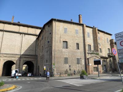 A large, historic brick building complex with arched ground-floor passageways, shuttered windows, and chimneys, viewed from a street corner with traffic signs (including one reading "Z.T.L." and "Comune di Parma") and a cyclist in the foreground.