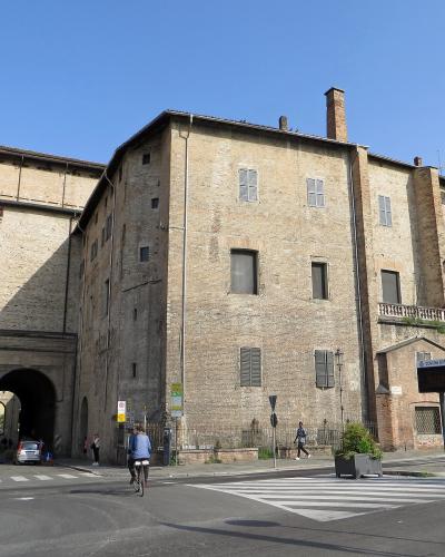 A large, historic brick building complex with arched ground-floor passageways, shuttered windows, and chimneys, viewed from a street corner with traffic signs (including one reading "Z.T.L." and "Comune di Parma") and a cyclist in the foreground.