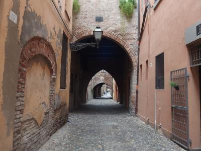 A narrow cobblestone street in Ferrara, Italy, viewed through a brick archway, with another arch further down the lane and buildings featuring shuttered windows and aged brickwork.