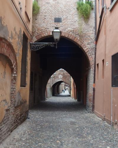 A narrow cobblestone street in Ferrara, Italy, viewed through a brick archway, with another arch further down the lane and buildings featuring shuttered windows and aged brickwork.
