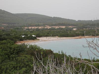 A coastal landscape showing a sandy beach curving along turquoise water, backed by dense green Mediterranean scrub and forested hills, with a few small buildings and people visible on the shore.