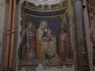 The image shows an interior fresco depicting the Madonna and Child enthroned with two angels, set within a painted architectural niche, flanked by brick columns and a stone column with a golden crucifix in the foreground.