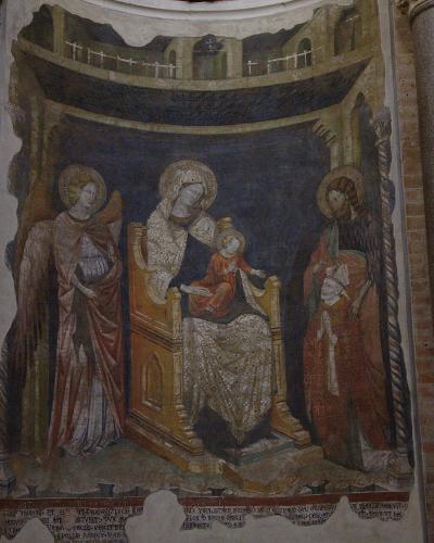 The image shows an interior fresco depicting the Madonna and Child enthroned with two angels, set within a painted architectural niche, flanked by brick columns and a stone column with a golden crucifix in the foreground.