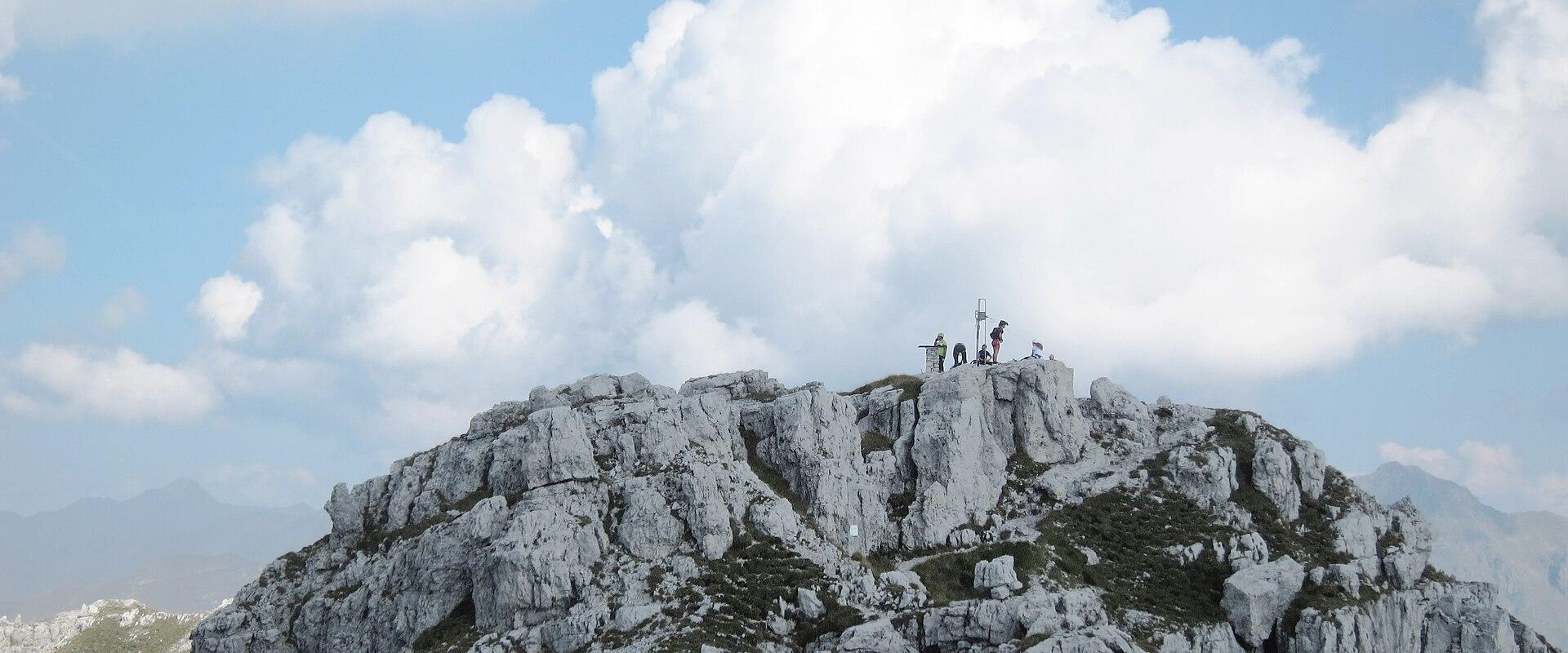 A rocky mountain summit with several people standing near a small stone structure and a metal pole, under a cloudy blue sky.