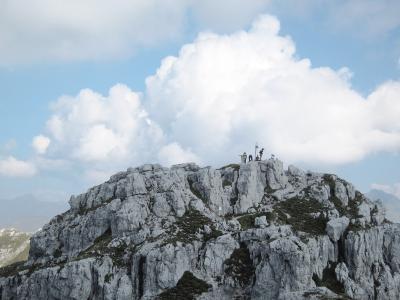 A rocky mountain summit with several people standing near a small stone structure and a metal pole, under a cloudy blue sky.