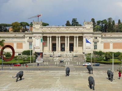 The image shows the exterior of the Galleria Nazionale d'Arte Moderna e Contemporanea in Rome, Italy, featuring its neoclassical facade, a large courtyard with lion sculptures, and a banner reading "TIME IS OUT OF JOINT".