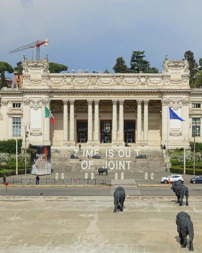 The image shows the exterior of the Galleria Nazionale d'Arte Moderna e Contemporanea in Rome, Italy, featuring its neoclassical facade, a large courtyard with lion sculptures, and a banner reading "TIME IS OUT OF JOINT".