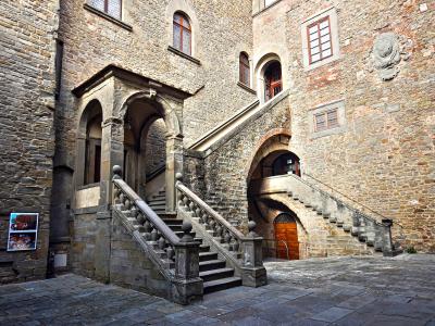 A stone courtyard featuring a prominent external staircase with balustrades, arched entryways, and a historic stone building facade with windows and a coat of arms.