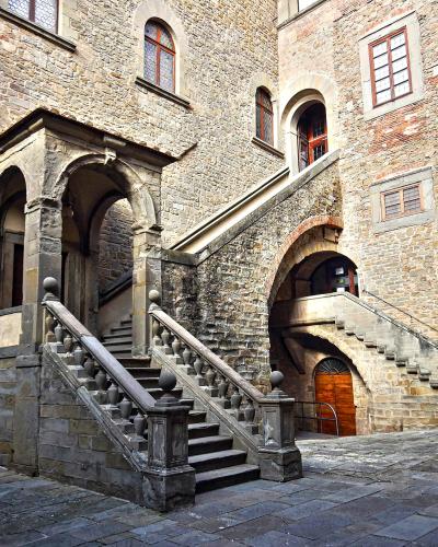 A stone courtyard featuring a prominent external staircase with balustrades, arched entryways, and a historic stone building facade with windows and a coat of arms.