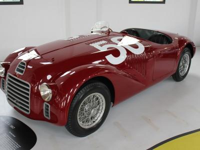 A red vintage Ferrari 125 S race car (number 58) displayed indoors on a polished floor with an informational plaque in the foreground.