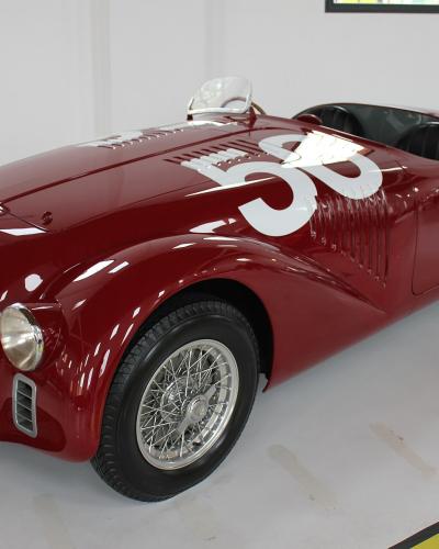 A red vintage Ferrari 125 S race car (number 58) displayed indoors on a polished floor with an informational plaque in the foreground.