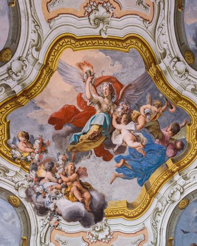 A Baroque-style ceiling fresco featuring a central winged female figure holding a torch, surrounded by cherubs and clouds, framed by ornate white and gold stucco moldings.
