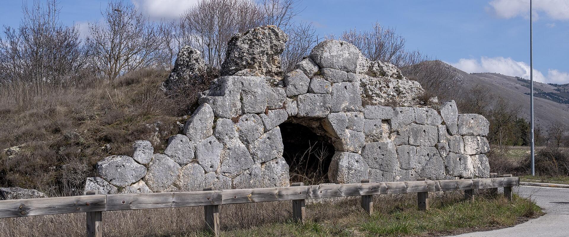 A stone ruin with an arched opening, situated beside a paved road and wooden guardrail, under a blue sky with scattered clouds; no visible signage or text.