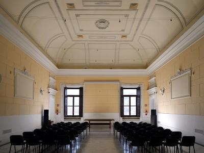 A large, empty hall with rows of black chairs facing forward, featuring a high ceiling with decorative plasterwork and two tall windows at the far end.