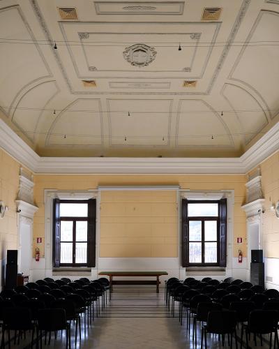 A large, empty hall with rows of black chairs facing forward, featuring a high ceiling with decorative plasterwork and two tall windows at the far end.