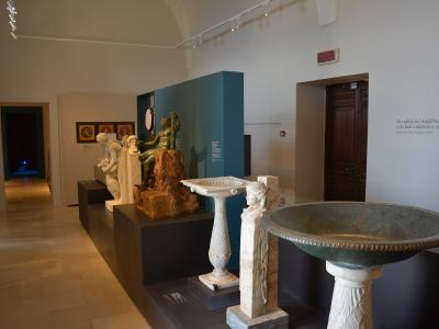 The image shows an interior museum gallery with classical sculptures, including a bronze statue on a rock base, marble busts, and stone basins, displayed on dark pedestals under track lighting, with informational text on the walls.