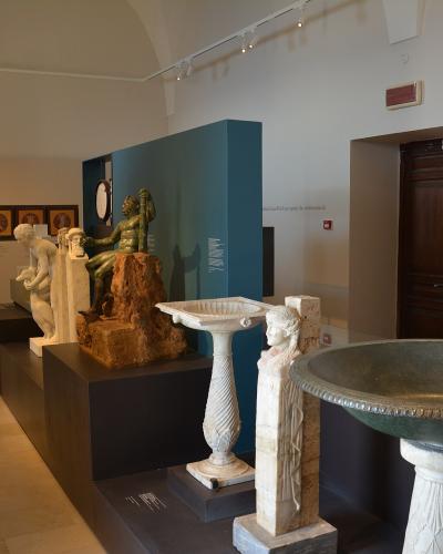 The image shows an interior museum gallery with classical sculptures, including a bronze statue on a rock base, marble busts, and stone basins, displayed on dark pedestals under track lighting, with informational text on the walls.