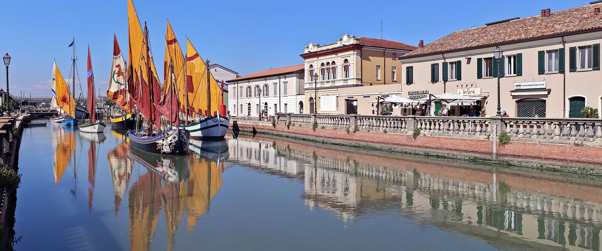 The image shows a canal lined with historic wooden sailing boats featuring tall yellow and red sails, moored along a stone quay with traditional Italian buildings in the background under a clear blue sky.