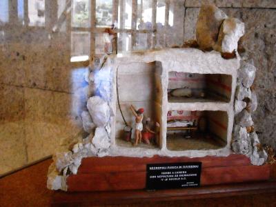 A scale model of a Punic tomb (tomba a camera) with painted interior details, displayed on a wooden base with a black plaque, inside a building with a window in the background.