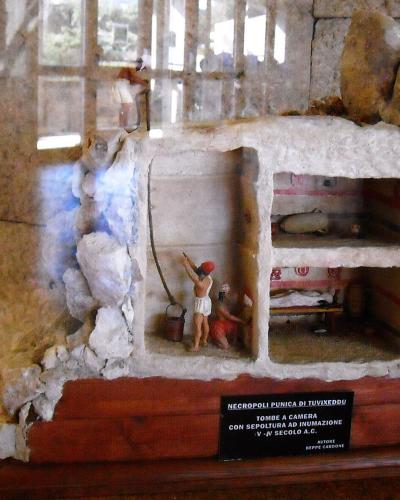 A scale model of a Punic tomb (tomba a camera) with painted interior details, displayed on a wooden base with a black plaque, inside a building with a window in the background.