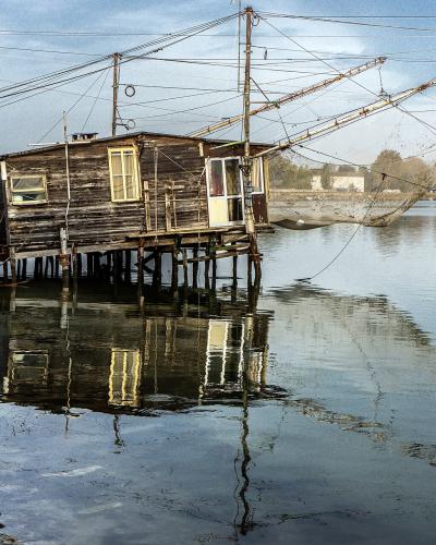 A weathered wooden fishing hut (cason) on stilts over calm water, featuring a large traditional fishing net structure extending from its roof, with a distant town skyline visible across the lagoon.