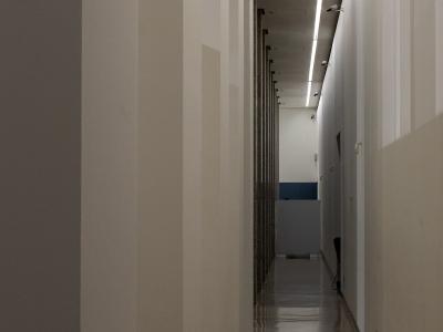 A narrow, modern interior hallway with white walls, a reflective floor, and a row of dark vertical panels or shelving units along the left side, illuminated by a long linear ceiling light.