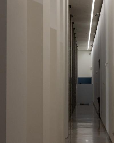 A narrow, modern interior hallway with white walls, a reflective floor, and a row of dark vertical panels or shelving units along the left side, illuminated by a long linear ceiling light.