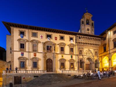 A large, illuminated historic palace facade at dusk featuring a prominent clock tower, multiple coats of arms, and an outdoor seating area in front.