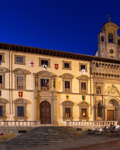 A large, illuminated historic palace facade at dusk featuring a prominent clock tower, multiple coats of arms, and an outdoor seating area in front.
