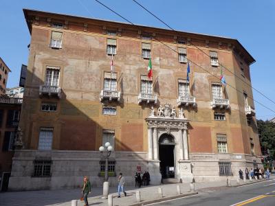 The image shows a large, historic building with a rusticated stone base and a frescoed upper facade, featuring a grand central entrance with statues and multiple flags (Italian, EU, and others), identified by the filename as Palazzo Doria Spinola,