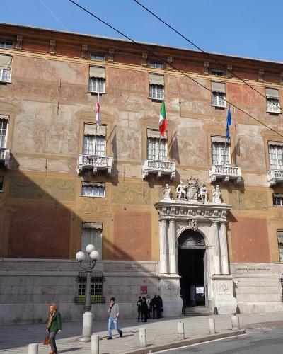 The image shows a large, historic building with a rusticated stone base and a frescoed upper facade, featuring a grand central entrance with statues and multiple flags (Italian, EU, and others), identified by the filename as Palazzo Doria Spinola,