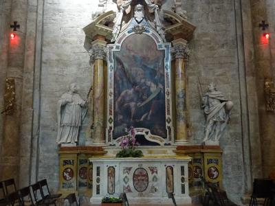 The image shows an ornate Baroque-style side altar inside a stone-walled church, featuring a central painting flanked by two statues, with a checkered floor and simple folding chairs in the foreground.