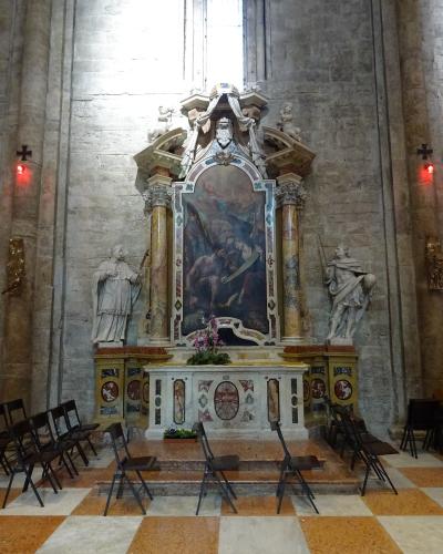 The image shows an ornate Baroque-style side altar inside a stone-walled church, featuring a central painting flanked by two statues, with a checkered floor and simple folding chairs in the foreground.