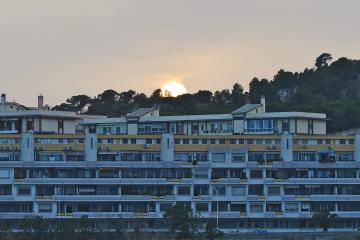 A large, multi-story residential apartment complex with yellow and grey facade sections, balconies, and air conditioning units, set against a hillside with trees and a setting sun.