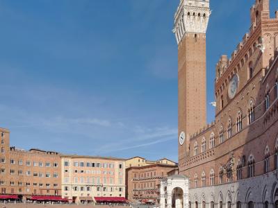 The image shows the Palazzo Pubblico and its tall Torre del Mangia tower in Siena, Italy, situated on the edge of the large, shell-shaped Piazza del Campo under a clear blue sky.