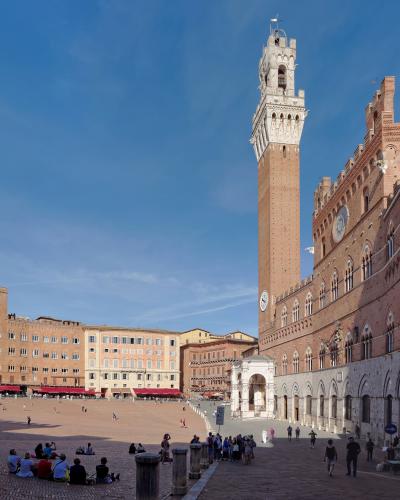 The image shows the Palazzo Pubblico and its tall Torre del Mangia tower in Siena, Italy, situated on the edge of the large, shell-shaped Piazza del Campo under a clear blue sky.