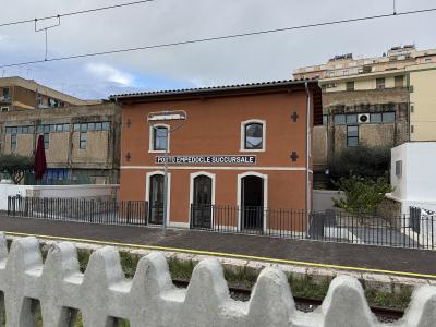 A two-story terracotta-colored building situated on a train platform, featuring a prominent black sign with white text reading "PORTO EMPEDOCLE SUCCURSALE" and railway tracks in the foreground.