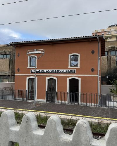 A two-story terracotta-colored building situated on a train platform, featuring a prominent black sign with white text reading "PORTO EMPEDOCLE SUCCURSALE" and railway tracks in the foreground.