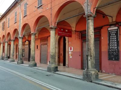 A street-level view of a long, terracotta-colored building with a covered arcade of stone columns and arches, featuring a red banner that reads "MUSEO DELLA RESISTENZA DI BOLOGNA" above an entrance.