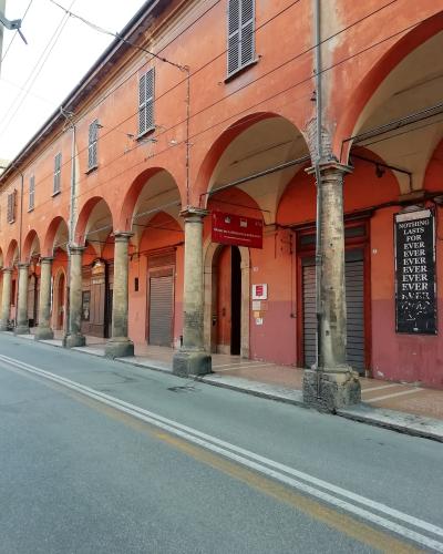 A street-level view of a long, terracotta-colored building with a covered arcade of stone columns and arches, featuring a red banner that reads "MUSEO DELLA RESISTENZA DI BOLOGNA" above an entrance.