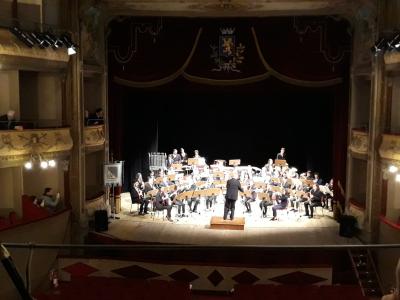 An interior view of a historic theater stage featuring an orchestra performing under a conductor, with a coat of arms above the proscenium arch and balconies on the left side.