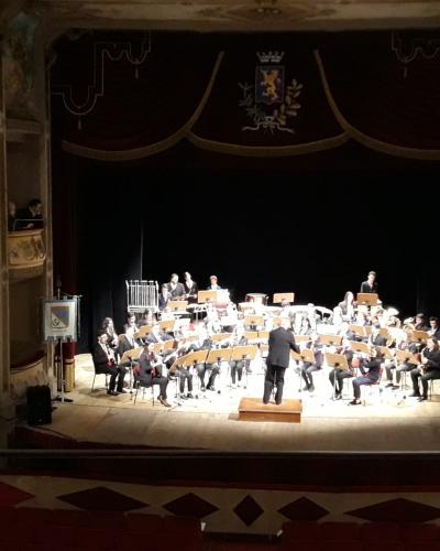 An interior view of a historic theater stage featuring an orchestra performing under a conductor, with a coat of arms above the proscenium arch and balconies on the left side.