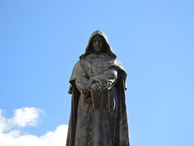 A bronze statue of a hooded figure holding a book, set against a blue sky with clouds, identified by the filename as Giordano Bruno in Campo de' Fiori.